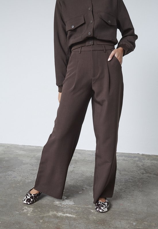 Model in een bruine broek met wijde pijpen en een bijpassend shirt met opgestikte zakken. Schoenen met koeienprint.