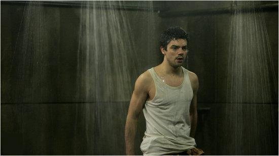 Een natte man in een witte tanktop staat onder een douche met waterstralen.