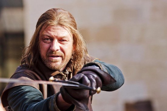 Man met lang bruin haar en baard, gekleed in bruin en groen, houdt een zwaard vast.