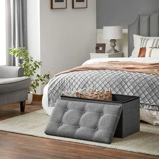 Grijze opbergkist met openklapbaar deksel met knopen, in een slaapkamer naast een bed.