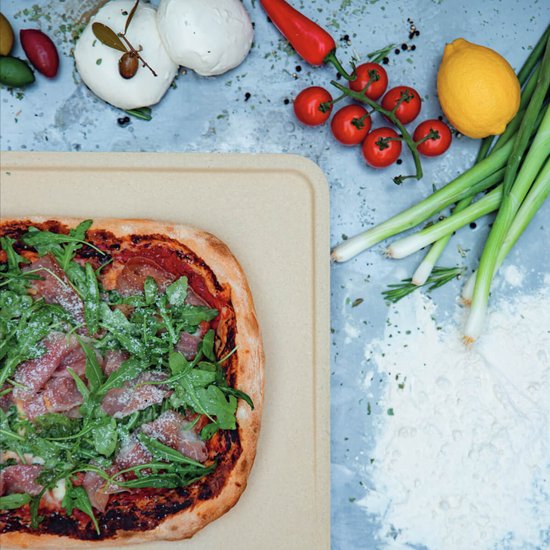 Pizza met ham, rucola en parmezaan op een beige pizzasteen, omringd door ingrediënten.
