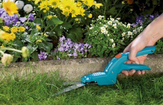 Hand met groen-blauwe Gardena grasschaar knipt gras naast een bloembed met gele en paarse bloemen.
