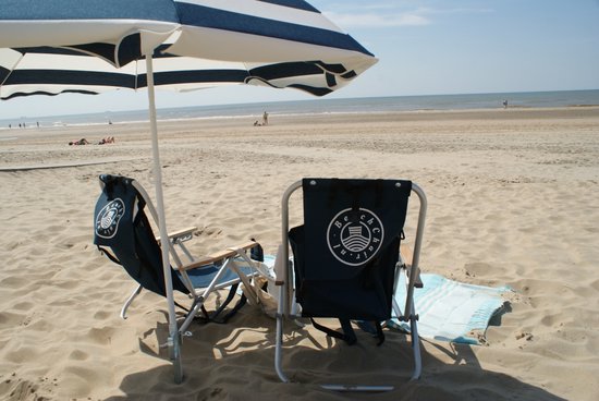 Twee donkerblauwe campingstoelen onder een gestreepte parasol op een zonnig strand.