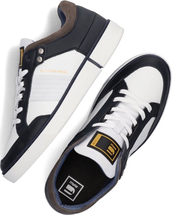 Set van twee witte sneakers met marineblauwe en bruine accenten. Eén sneaker toont de zool met 'RAW' logo.