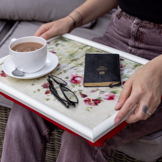 Vrouw met dienblad op schoot. Het dienblad heeft een bloemenprint van rode en witte klaprozen. Naast een kopje koffie liggen een bril en een boekje.