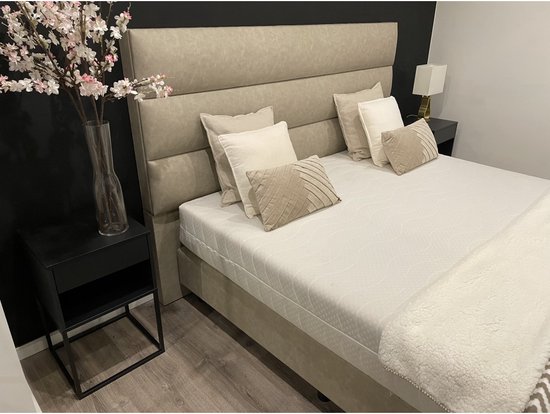 Beige boxspring met gestoffeerd hoofdbord, luxe kussens en beige deken met geometrisch patroon.