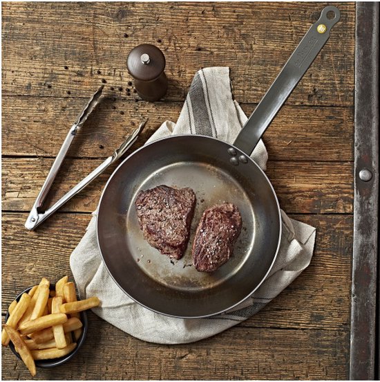 Zilveren koekenpan met twee stukken steak erin, op een houten tafel. Naast de pan liggen frietjes en een pepermolen.