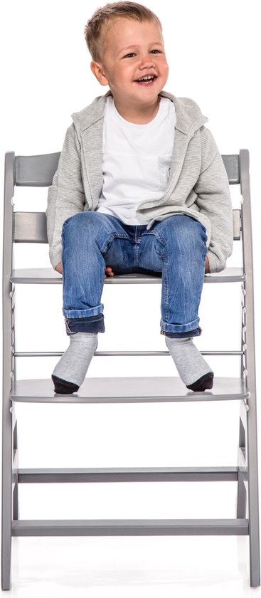 Een jongen met een grijze hoodie, wit shirt en blauwe jeans zit lachend op een grijze kinderstoel.
