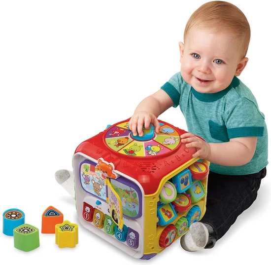 Een baby in een groen T-shirt speelt met een veelkleurige VTech activiteitenkubus met dierengeluiden en knoppen.