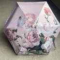 Zespanelen paraplu, gesloten, met een fel roze pioenrozen schilderijprint op een lichtere roze achtergrond.