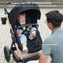 Jongetje met blauwe jeans zit in een zwarte kinderwagen met 5-puntsgordel, kijkt naar man.