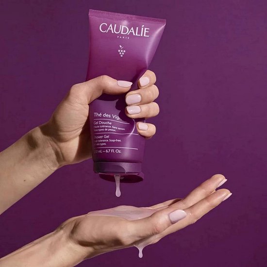 Een hand met lichte nagellak houdt een Caudalie douchegel tube vast, terwijl de douchegel in de andere hand druppelt.