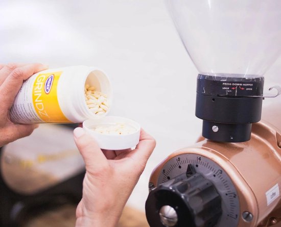 Een witte pot met gele dop, gevuld met kleine witte pellets, die naast een koffiemolen wordt gehouden.