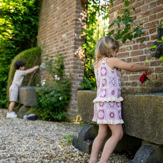 Meisje met blonde vlechtjes, lila top met bloemenprint en ruches, bij een stenen fontein.
