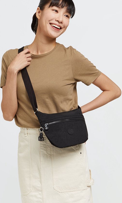 Vrouw met zwarte Kipling Arto S schoudertas, model met zilverkleurige ritsen en aapje. Ze draagt een bruin T-shirt en een crème rok.