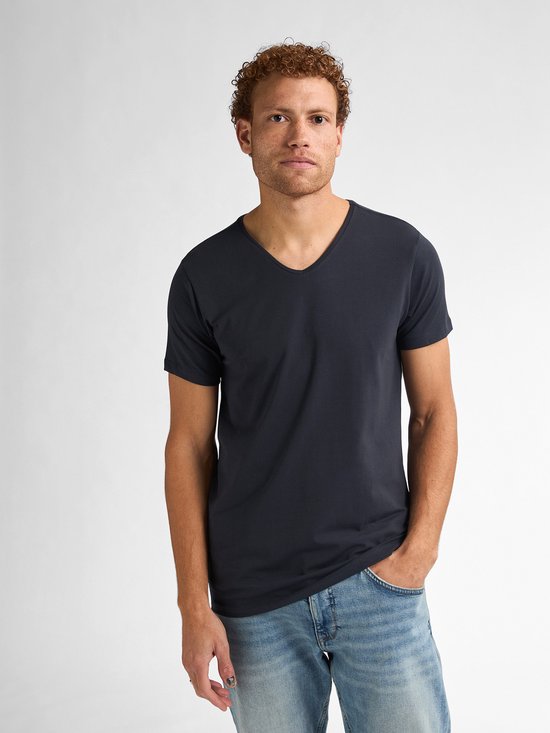 Man met krullend bruin haar draagt een donkerblauwe V-hals T-shirt en een spijkerbroek.