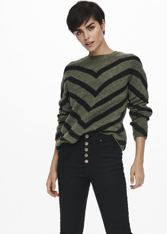 Model in olijfgroene trui met zwarte V-strepen en zwarte broek.