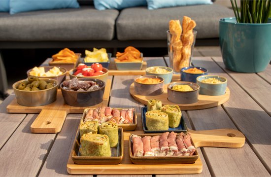Een gedekte tafel met diverse hapjes op bamboe serveerplanken. De planken bevatten onder andere rolletjes, ham en dips.