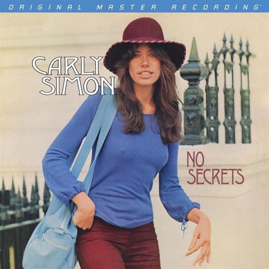 Carly Simon draagt een bordeauxrode hoed, blauwe top en bruine broek, met een blauwe tas over haar schouder.