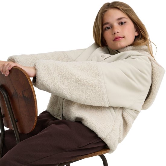 Meisje op een stoel in beige jas met teddy-inzet op de mouwen en zakken, en capuchon.