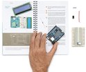 Een hand houdt een Arduino-bord vast boven een open boek met instructies, met elektronische componenten op een tafel.