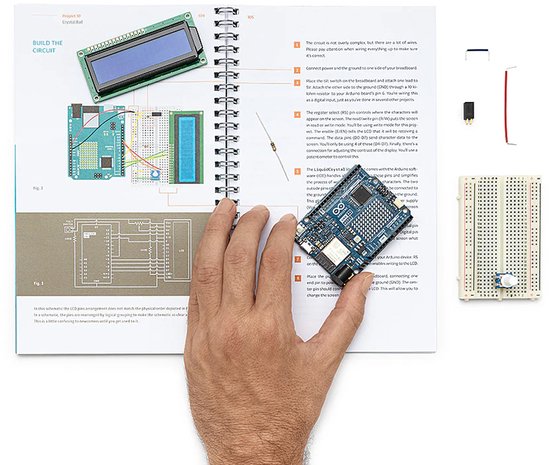 Een hand houdt een Arduino-bord vast boven een open boek met instructies, met elektronische componenten op een tafel.