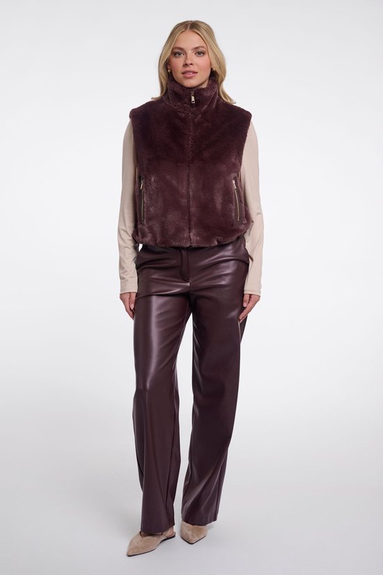 Model in bruin kort gilet met rits en hoge kraag, beige coltrui en bruine leren broek, staand.
