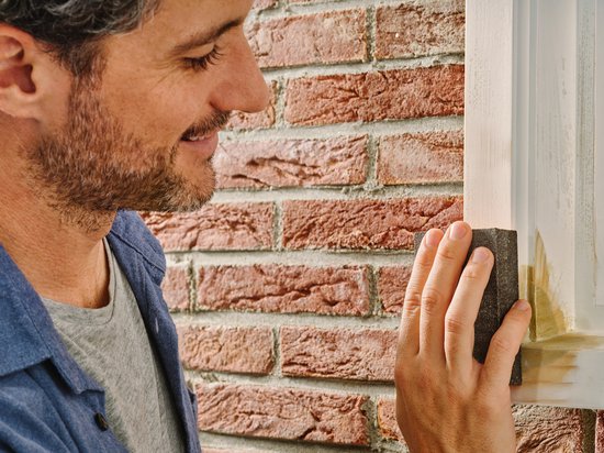 Een man schuurt met een schuurblok een reparatie aan een wit kozijn naast een bakstenen muur.