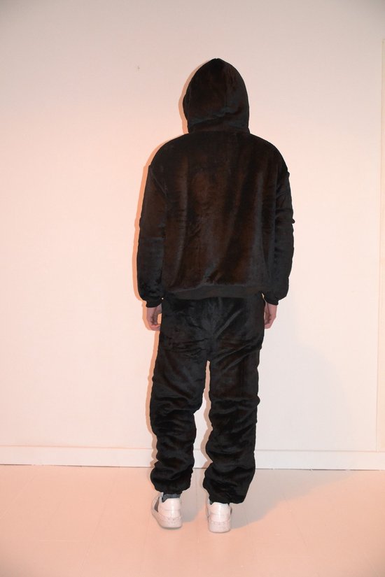 De achterkant van een oversized zwart trainingspak met capuchon en een rood S-logo op de linkerbroekspijp, gedragen door een man. Het pak is gemaakt van zacht materiaal en de broek is gerimpeld aan de onderkant. Hij draagt witte sneakers.