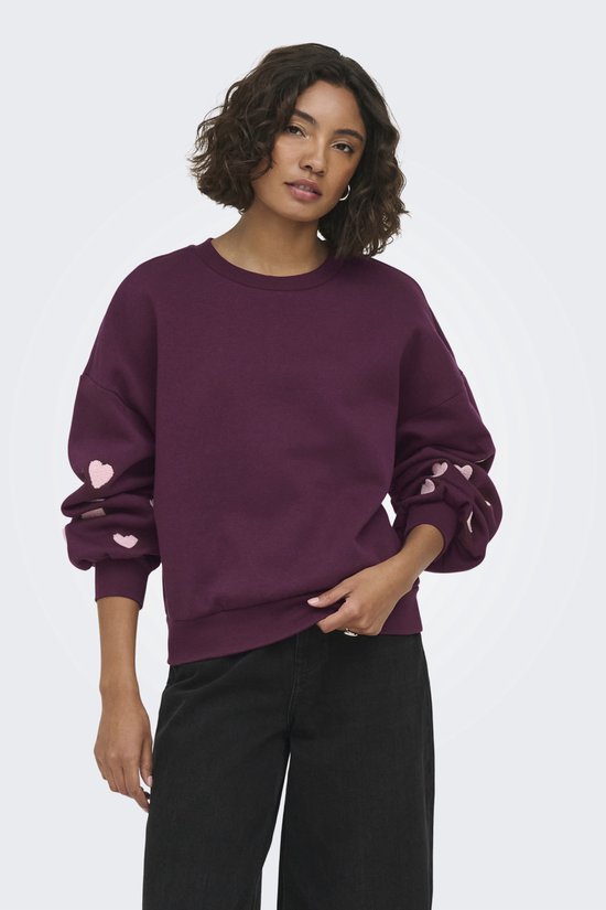 Model draagt een donkerpaarse sweater met lichtroze hartjes op de mouwen en een zwarte broek.