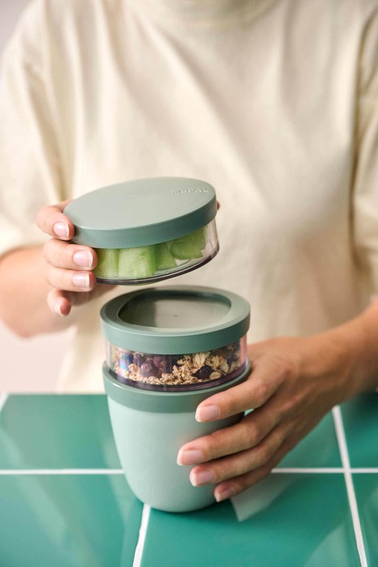 Een persoon demonteert een groene lunchpot met twee compartimenten, gevuld met muesli en stukjes meloen.