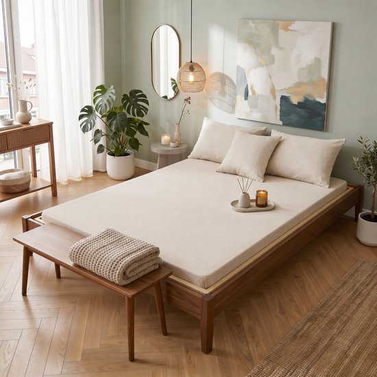 Un lit double fait avec une housse de couette beige clair, des oreillers et un plaid. La pièce a des plantes et de la lumière naturelle.