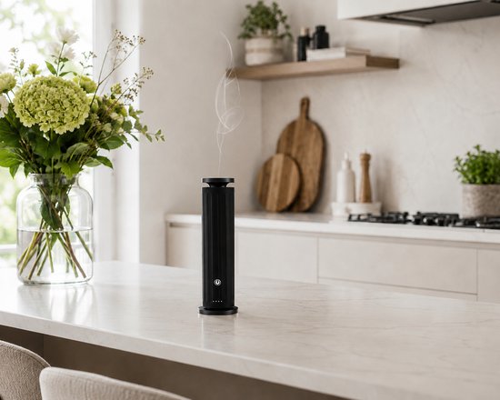 Zwarte, verticale aroma diffuser op een wit aanrecht, met dunne rookpluimen die eruit komen. Op de achtergrond een vaas met bloemen.