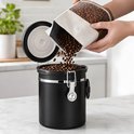 Een hand die koffiebonen uit een zak stort in de open zwarte RVS koffiebewaarpot.