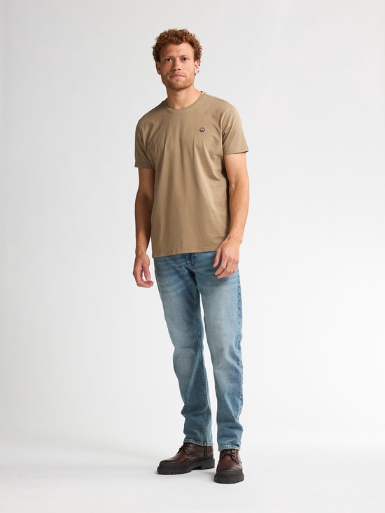 Man met krullend rood haar, staat frontaal, draagt lichtbruin t-shirt met borstlogo en spijkerbroek.