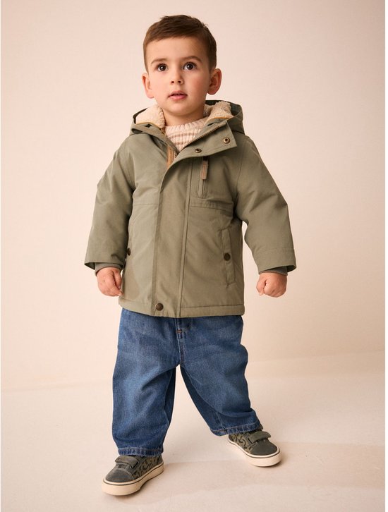Een jongetje in een groene parka met capuchon, spijkerbroek en sneakers, staand tegen een neutrale achtergrond.