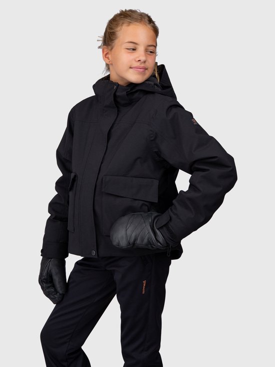 Meisje in zwarte ski-jas met capuchon, ski-broek en ski-handschoenen, hand in de zij.