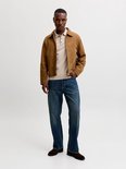 Man met beige gebreid poloshirt en bruine suède jas, met donkerblauwe jeans.