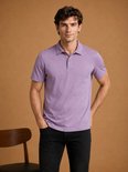 Een man met donker haar draagt een lavendelkleurig poloshirt met korte mouwen en zwarte jeans, handen in de zakken.