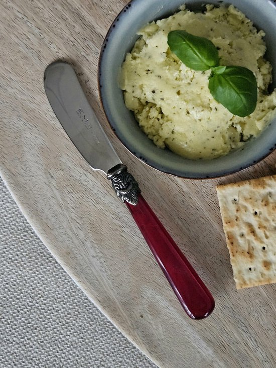 Boter mesje met zilverkleurig sierlijk handvat, rood handvat, naast een kommetje kruidenboter en een cracker.