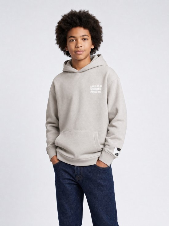 Jongen draagt een lichtgrijze hoodie met kleine witte tekst op de borst en donkere jeans. Hij heeft handen in zijn zakken.