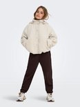 Meisje staat met handen in de zakken van een beige teddy jas, bruine joggingbroek.