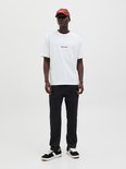 Man met witte t-shirt, zwarte joggingbroek met geborduurde tekst en bruine pet.