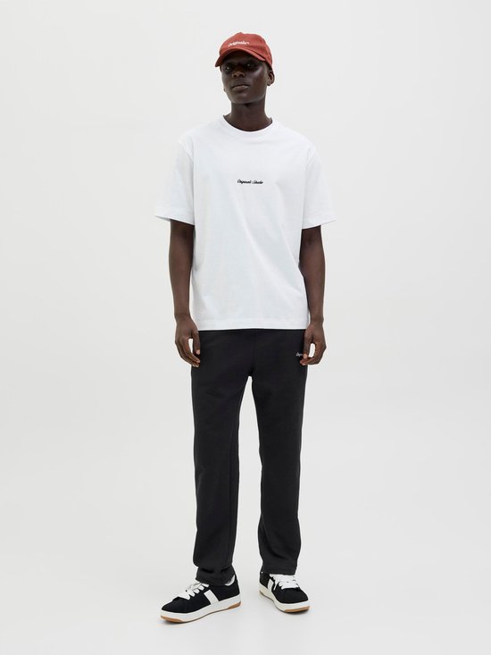 Man met witte t-shirt, zwarte joggingbroek met geborduurde tekst en bruine pet.