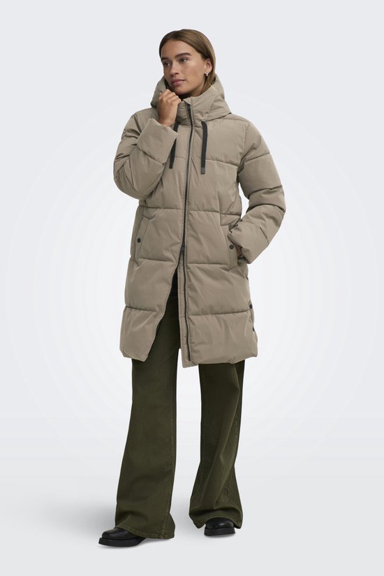 Vrouw in lang, beige pufferjack met capuchon, de rits gedeeltelijk gesloten en handen in de zakken.