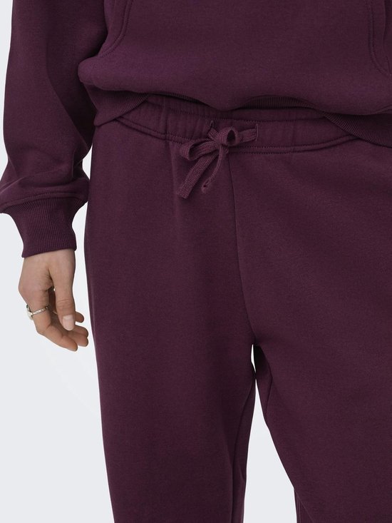 Detail van de bordeauxrode joggingbroek met koordje in de taille en een zilverkleurige ring aan de hand.