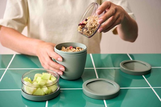 Handen vullen een groen Mepal lunchpot met muesli. Naast de pot ligt een ander potje met stukjes meloen.