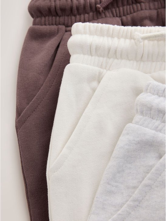 Drie joggingbroeken, close-up van de taillebanden met trekkoorden en steekzakken in bruin, gebroken wit en lichtgrijs.