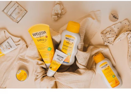 WELEDA Baby producten met oranje en gele accenten, waaronder een tube shampoo, een tube billenbalsem en een flesje verzorgende olie.