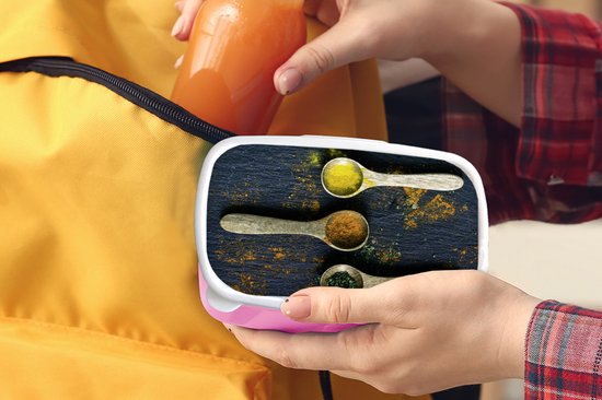 Boîte à pain rose avec couvercle représentant des épices sur des cuillères, glissée dans un sac jaune.